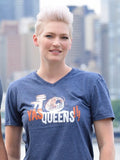 Woman wearing a blue t-shirt with 'YAS QUEENS!!!' text and graphic, standing outdoors with a cityscape background.