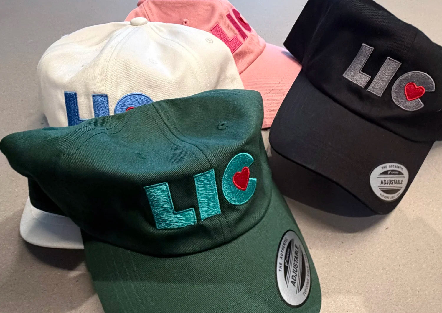 Four baseball caps in pink, white, black, and green colors with 'LIC' colored branding