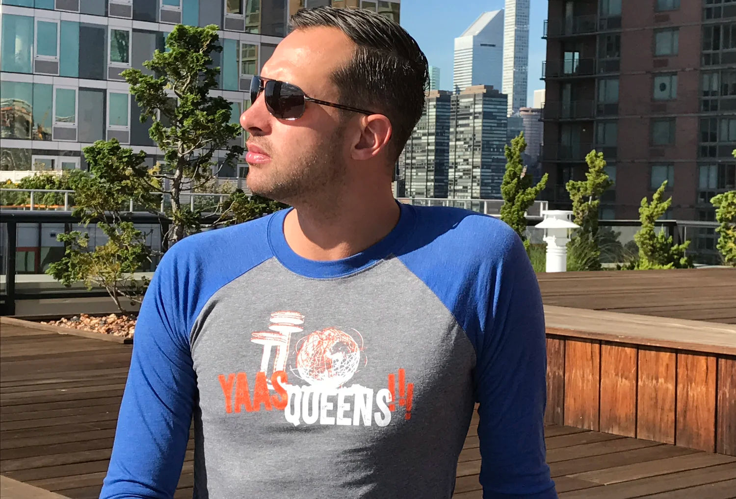 Man wearing a gray and blue long-sleeve shirt with 'YAAS QUEENS' graphic text on a rooftop with cityscape background