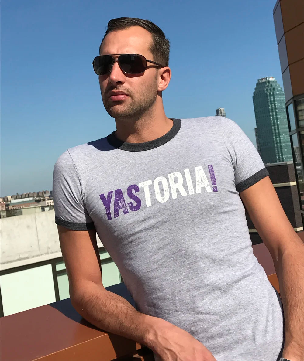 Man wearing a gray t-shirt with 'YASTORIA' printed on it, standing on a rooftop with a cityscape background.