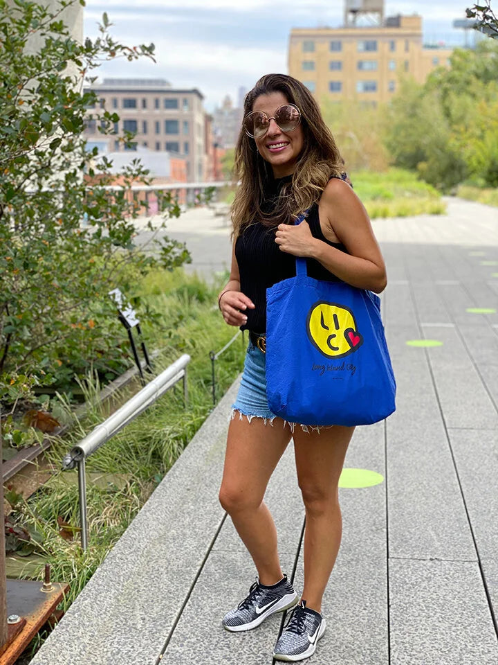 Woman carrying a blue tote bag with a smiley face design outdoors.