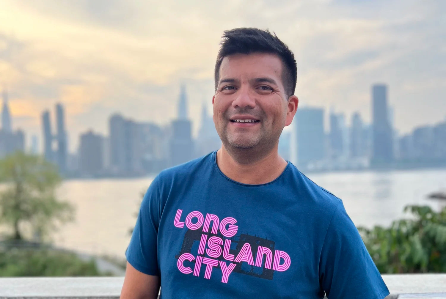 Man wearing a blue t-shirt with 'Long Island City' neon graphic text, standing in front of a city skyline.