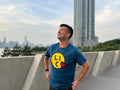 Smiling man wearing a blue t-shirt with a smiley face design, standing on a concrete ledge with a cityscape in the background.