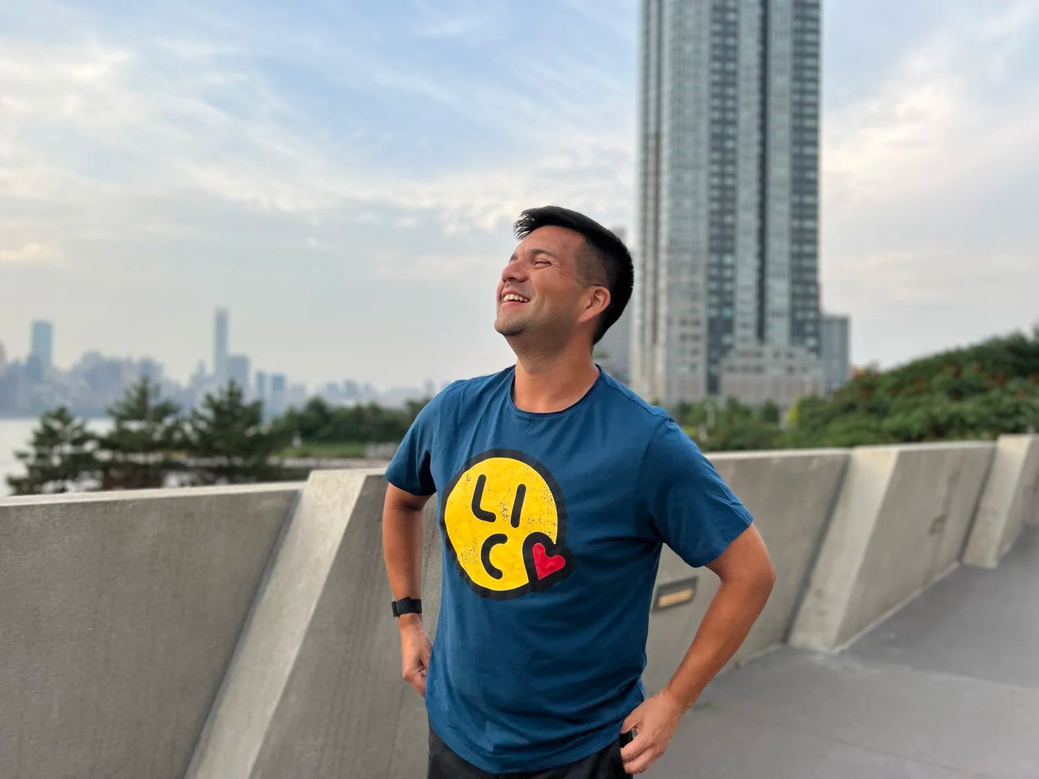 Smiling man wearing a blue t-shirt with a smiley face design, standing on a concrete ledge with a cityscape in the background.