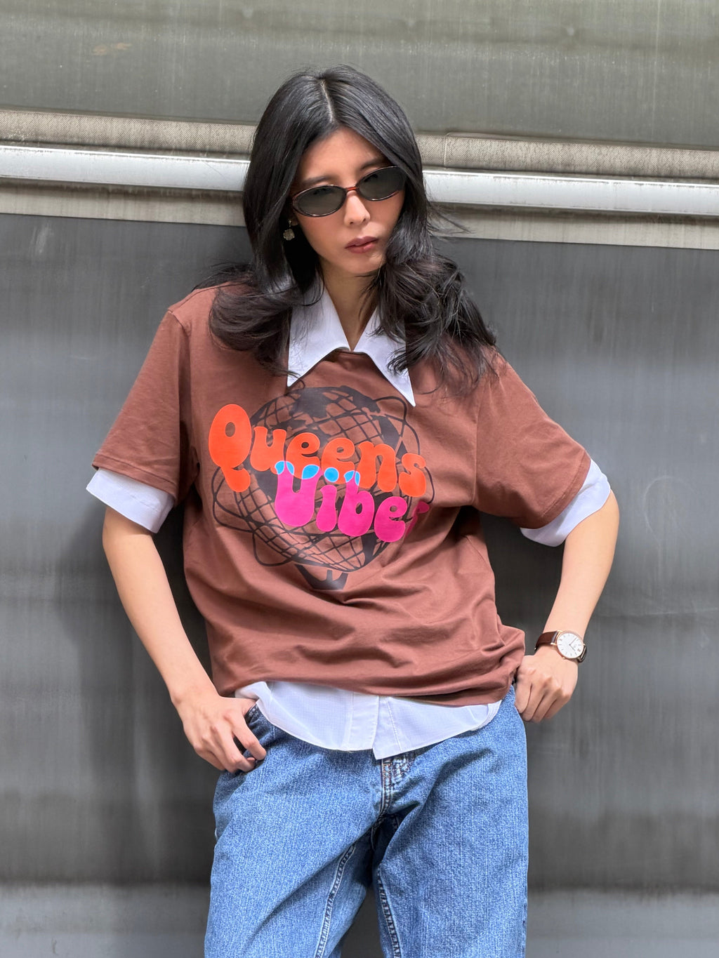 Person wearing a brown t-shirt with 'Queens Wife' text and blue jeans against a metallic wall.