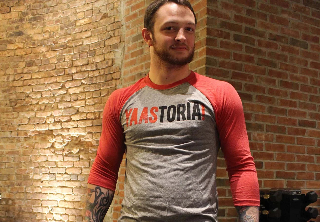 Man wearing a red and gray long-sleeve shirt with 'YAASTORIA' text against a brick wall.