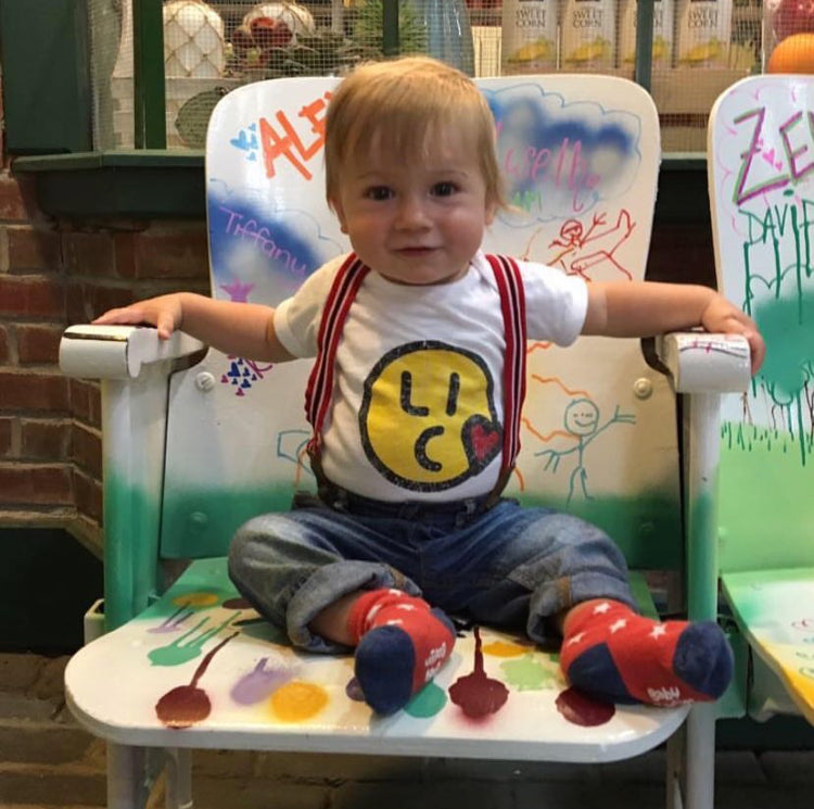 Toddler wearing smiley face logo white t-shirt sitting in a colorful high chair with various drawings and letters on the backrest.