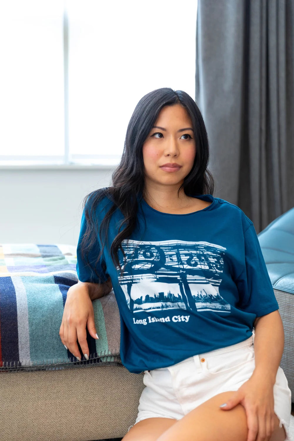 Woman wearing a blue t-shirt with a Long Island City text with graphic design, sitting on a couch.