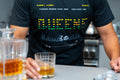Person wearing a black t-shirt with a 'QUEENS' digital graphic text holding a glass of whiskey