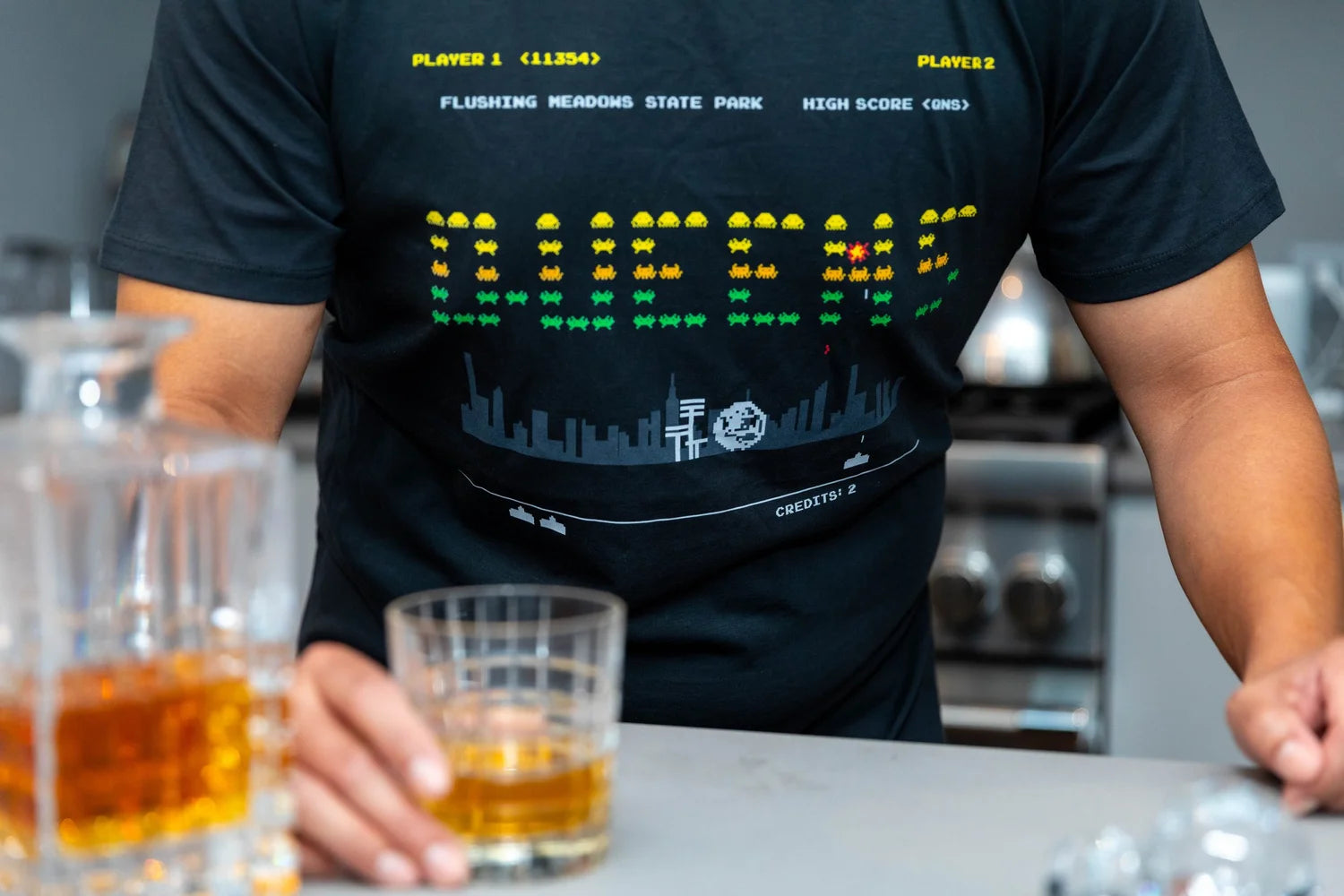 Person wearing a black t-shirt with a 'QUEENS' digital graphic text holding a glass of whiskey