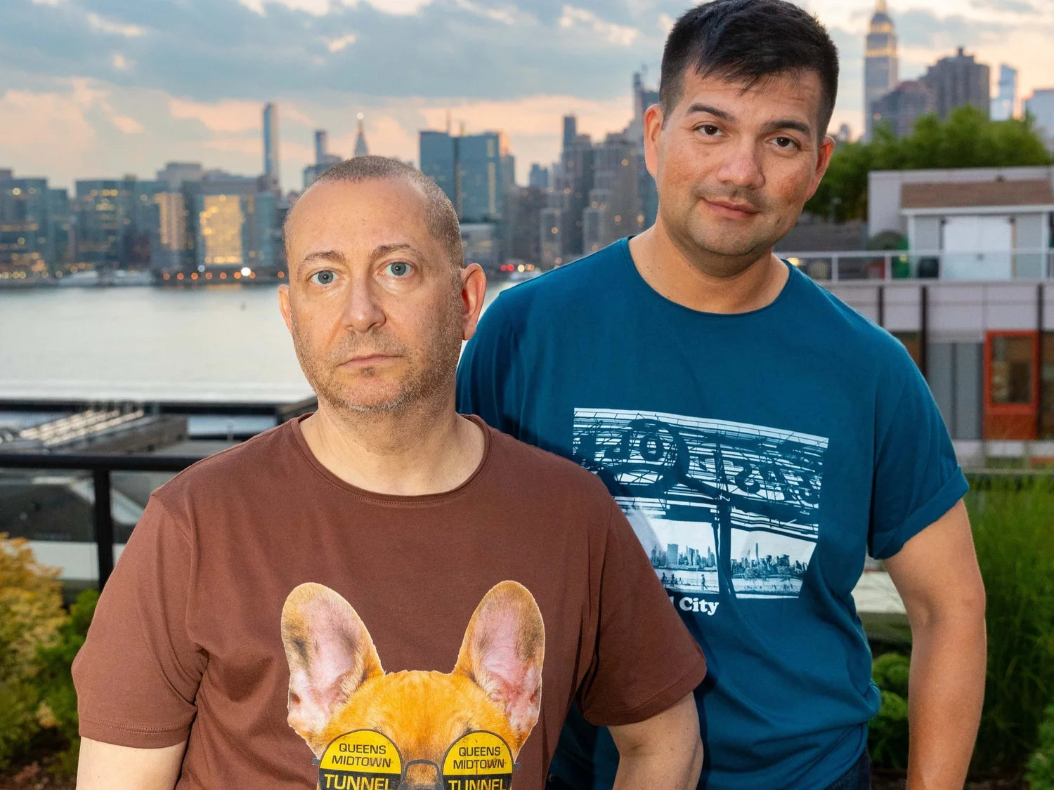Two men standing outdoors with a city skyline in the background wearing a brown and dark blue t-shirt featuring Long Island City graphics