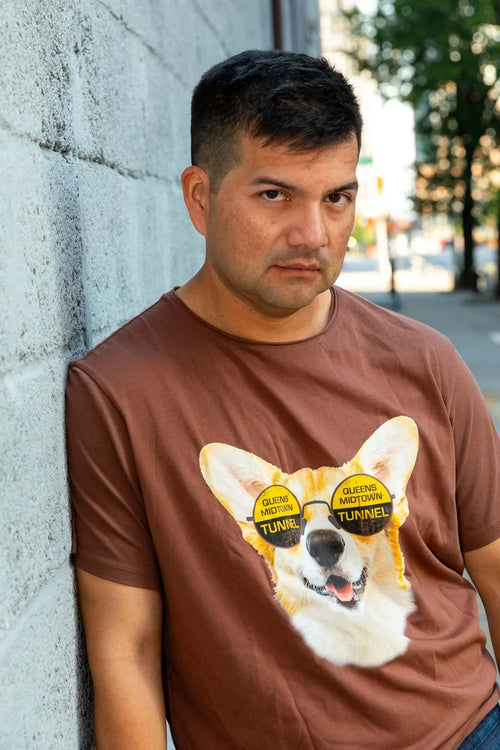 Man wearing a brown t-shirt with a dog with 'Queens Midtown Tunnel' text glasses graphic leaning against a wall.