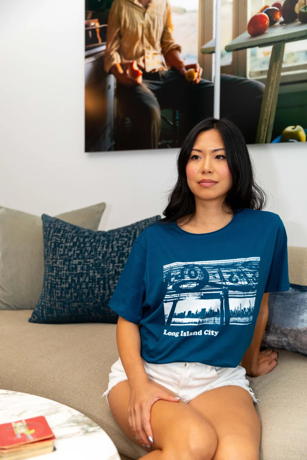 Woman wearing a blue t-shirt with a Long Island City text with graphic design, sitting on a couch.