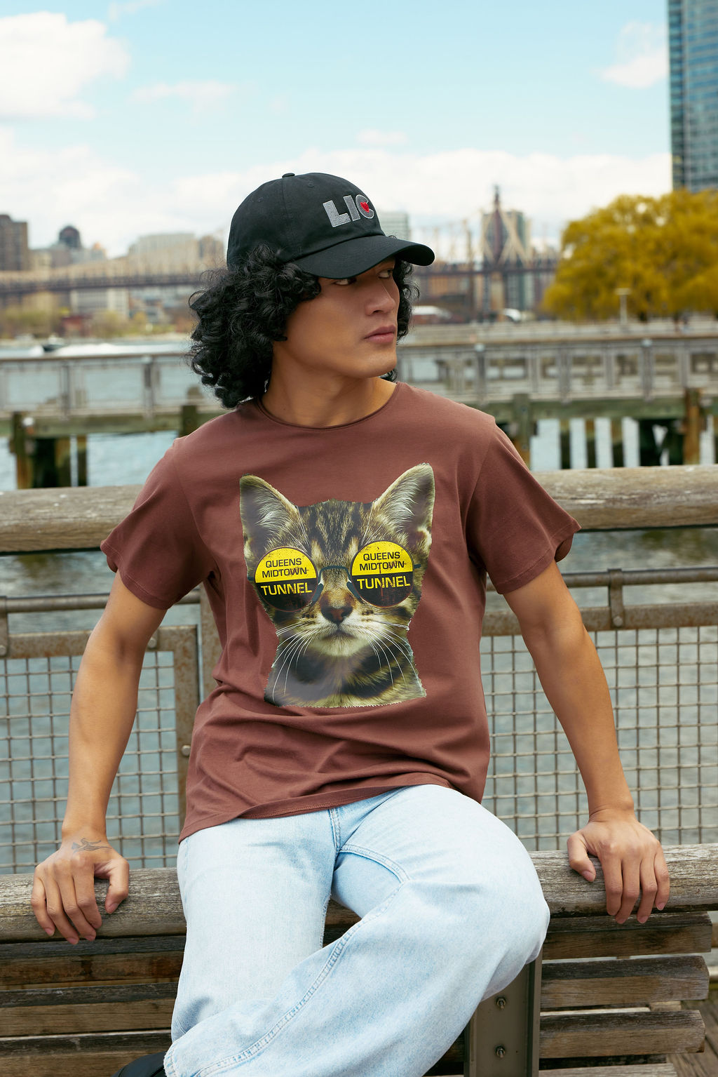 Person wearing a brown t-shirt with a cat graphic and text, sitting on a wooden bench by a waterfront with a cityscape in the background.
