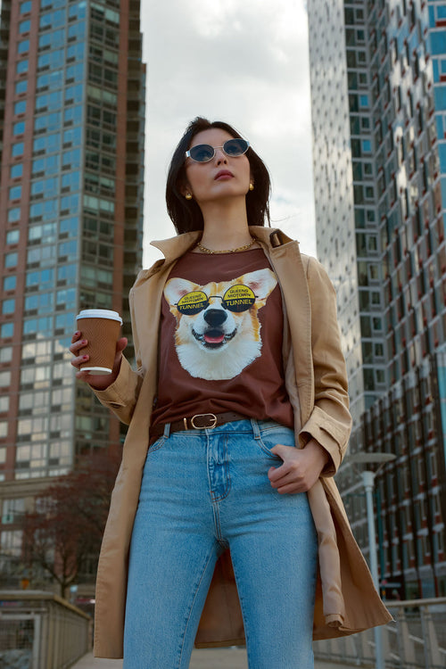 Woman wearing a brown t-shirt with a dog graphic, blue jeans, and sunglasses, holding a coffee cup in an urban setting.