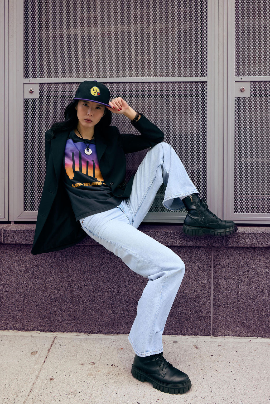 Person wearing a black hoodie with a colorful graphic, light blue jeans, and black boots sitting on a ledge.