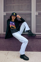 Person wearing a black hoodie with a colorful graphic, light blue jeans, and black boots sitting on a ledge.