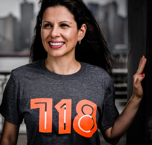 Woman wearing a dark gray t-shirt with '718' printed in orange, standing against an urban background.