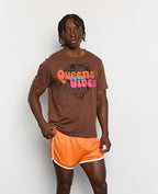 Person wearing a brown t-shirt with 'Queens vibes' text and orange shorts against a white background