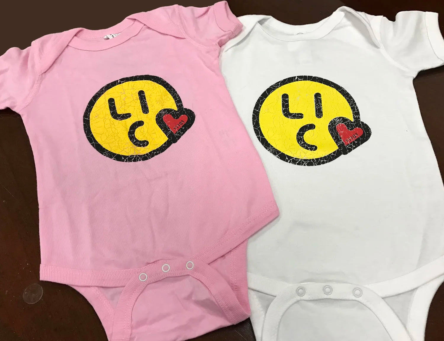 Pink and white baby onesies with smiley face "Long Island City" logo designs on a dark surface